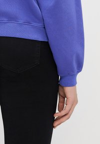 Sudadera púrpura con puños y dobladillo de canalé, combinada con jeans negros. La mano descansa suavemente sobre la pierna, mostrando las texturas de la tela.