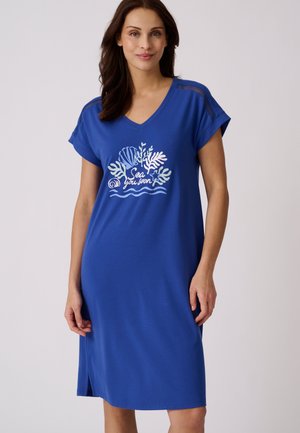 Vrouw met een blauwe jurk tot op de knie, korte mouwen en een schelpenontwerp met de tekst "Sea you soon" aan de voorkant.