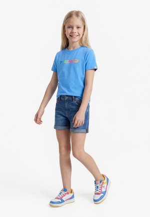 Niña sonriente con cabello rubio largo, vestida con una camiseta azul de manga corta, pantalones cortos de mezclilla y zapatillas coloridas, posando sobre un fondo blanco.