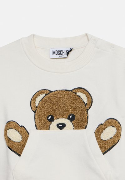 Sweat-shirt bébé couleur crème avec un motif d'ours en peluche marron partiellement glissé dans la poche avant et des boutons pression à l'épaule.