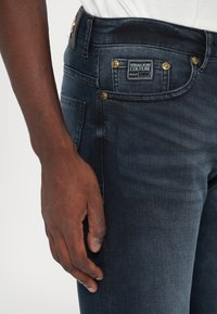 Σκούρα μπλε τζιν παντελόνια χαρακτηρίζονται από εφαρμοστή σχεδίαση με χρυσές λεπτομέρειες, συμπεριλαμβανομένων κουμπιών και μιας μαύρης ετικέτας στην πίσω τσέπη.