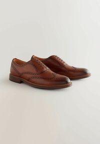 Pantofi brogue din piele maro, cu vârf rotund, design cu șiret, perforații intricate și un capac pe vârf de culoare închisă. Textură netedă.