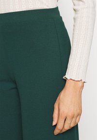 Gröna högmidjade byxor med texturerad yta, tillsammans med en vit långärmad topp med ribbat mönster och scallopade ärmslut.