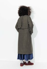 Manteau long en pied de poule marron avec un dos superposé, associé à une jupe en satin bleu marine et des baskets bordeaux. Tissu texturé et coupe ample.