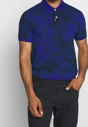 Homme portant un polo à manches courtes bleu foncé à motifs et un pantalon noir, une main dans la poche et l'autre détendue.