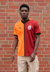 Puma fodboldtrøje har et delt design i orange og rød med sorte accenter. Indeholder et mærke med stjerner og et rundt logo på brystet.