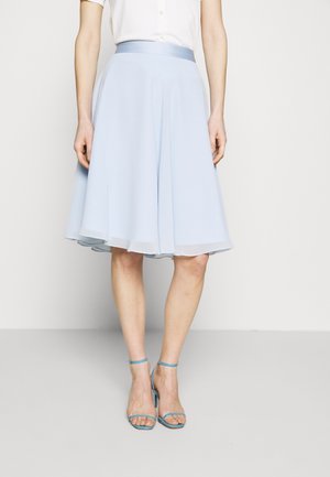 Mujer con falda azul claro acampanada hasta la rodilla, blusa blanca abotonada y sandalias de tacón alto con correa en el tobillo, sobre un fondo liso.