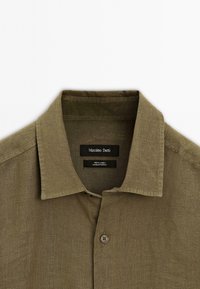 Olive green linen shirt featuring a classic collar, button closure, and a textured fabric. Label includes brand and fabric information.