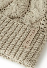 Beige stickad mössa med flätmönster, ribbad mudd och en läderetikett präglad med "FALCONER." Texturerat material med lätt glans.