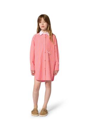 Jeune fille aux longs cheveux portant une robe-chemise rayée rose et orange surdimensionnée et des chaussures marron à enfiler, debout sur fond blanc.