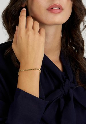 Bracciale d'oro con perle rotonde, indossato a una mano. Presenta una texture liscia e un design delicato. Modella che indossa un top scuro.