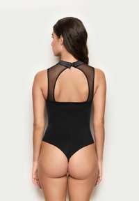 Kobieta w czarnym bezrękawnikowym body z wysokim dekoltem i przezroczystą siateczkową tylną częścią, stojąca tyłem do aparatu.