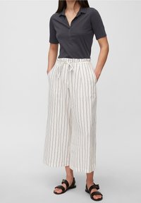 Kvinna som bär en kortärmad mörk polo t-shirt, vita högmidjade vida randiga byxor med bälte och svarta sandaler.