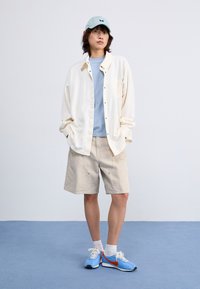 Svijetlosmeđa oversized košulja, plava majica, svijetlosmeđi šortsevi i plave tenisice s narančastim detaljima. U complet outfit-a nalazi se i podstavljena kapa.