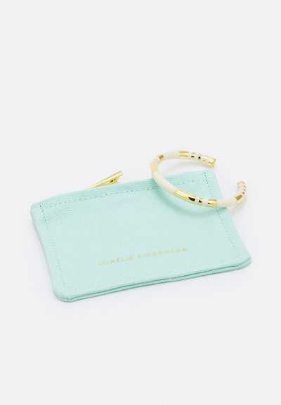 Turquoise canvas pouch with gold logo and zipper, accompanied by a gold-tone bracelet with white and striped detailing.