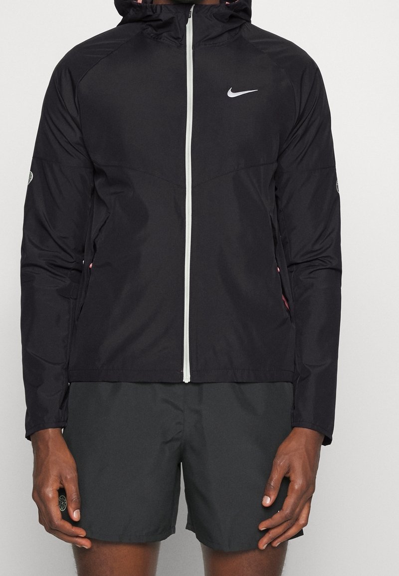 Mężczyzna w czarnej sportowej kurtce z podwójnym zamkiem na plecach Nike i czarnych szortach sportowych, stojący z ramionami w luźnej pozycji na tle jednolitym.