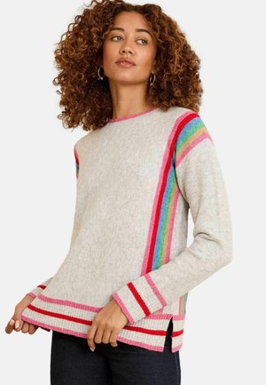Frau mit lockigem Haar, die einen hellgrauen Pullover mit roten, rosa, blauen, grünen und regenbogenfarbenen Streifen trägt, hält den Saum und schaut leicht nach rechts.