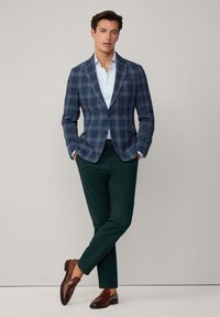 Námornícky károvaný blazer, svetlomodrá košeľa, tmavozelené nohavice, hnedé mokasíny. Blazer má klopové reverzy a jedno gombíkové zapínanie.