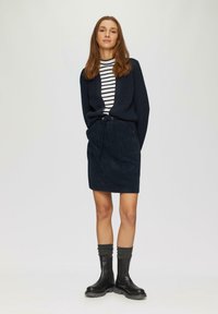 Marineblauer Strickcardigan über einem gestreiften Rollkragenpullover, kombiniert mit einem dunklen Cordrock und schwarzen Stiefeletten mit strukturierten grauen Socken.