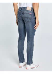 Személy kék karcsúsított farmernadrágban és fehér, piros és fekete részletekkel díszített sportcipőben, egyszerű világos háttér előtt állva.