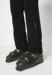 Black ski pants with a fitted design, paired with black ski boots featuring metal buckles and a textured finish. CMP logo on pants.