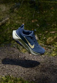 Blå Nike löparskor med beige swoosh i luften ovanför en grus- och gräsmark i utomhusmiljö.