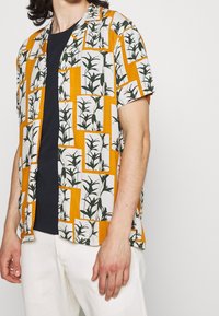 Kurzärmliges Hemd mit einem weißen und orangefarbenen geometrischen Muster sowie grünen Pflanzen-Silhouetten. Hergestellt aus leichtem Material.