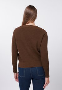 Καφέ πλεκτό πουλόβερ με ριμπ ύφανση, στρογγυλή λαιμόκοψη, μακριά μανίκια και ελαφρώς cropped τελείωμα, συνδυασμένο με μπλε τζιν παντελόνι.