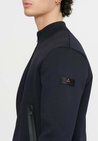 Giacca a zip color navy realizzata in tessuto testurizzato. Presenta un colletto alto, tasche laterali con zip e un'etichetta nera con punti rossi sulla manica.