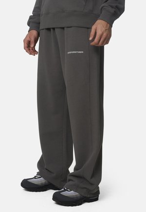 Uomo che indossa pantaloni della tuta ampi di colore grigio scuro con il testo "GOODFORNOTHING" sulla coscia sinistra e sneakers nere e grigie.