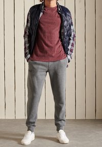 Navy gepolsterte Weste über einem karierten Langarmhemd, weinrotem strukturiertem T-Shirt, grauen Jogginghosen und weißen Sneakers. Holzwand-Hintergrund.