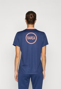 Navy athletic shirt with a slim fit, features a pink collar and circular "BARÇA" logo on the back in contrasting color. Smooth fabric texture.