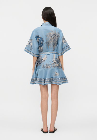 Γυναίκα που φοράει μπλε, κοντό φόρεμα με φαρδιά μανίκια και τροπικά και λουλουδένια σχέδια, στέκεται με την πλάτη στην κάμερα και μαύρα σανδάλια.