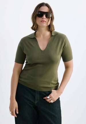 Femme portant des lunettes de soleil noires, un haut à manches courtes vert olive avec col, un jean foncé et des bagues argentées, posant avec une main dans la poche.