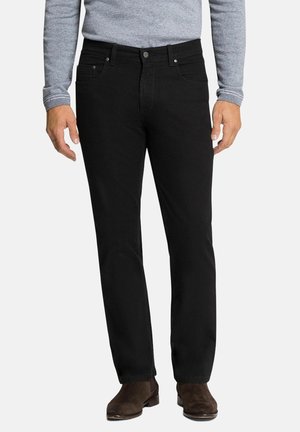 Man draagt zwarte broek met rechte pijpen, grijs shirt met lange mouwen en bruine suède enkellaarsjes, staand voor een effen achtergrond.