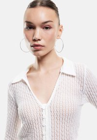 Weißer, strukturierter Cardigan mit kragenförmigem Ausschnitt, Knopfverschluss und einem Spitzenmuster. Das Model trägt große silberne Creolen.