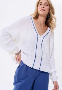Witte gebreide trui met een V-hals en blauwe accenten, met losse mouwen en een relaxed fit, gecombineerd met blauwe broek.