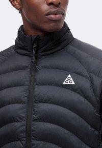 Gros plan d'une personne portant une doudoune noire à col montant avec un logo ACG triangulaire blanc sur le côté gauche de la poitrine.