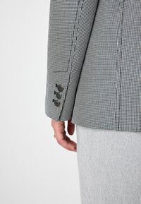 Zwart-wit houndstooth blazer met drie knopen op de manchet, textuurstof en een getailleerde pasvorm, getoond vanaf de achterkant met een grijze rok.