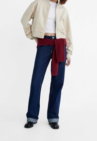 Beige ruskind bomberjakke, hvid crop top, mørkeblå bredbenede jeans med opsmøgede kanter og en rød strikket sweater bundet om taljen.