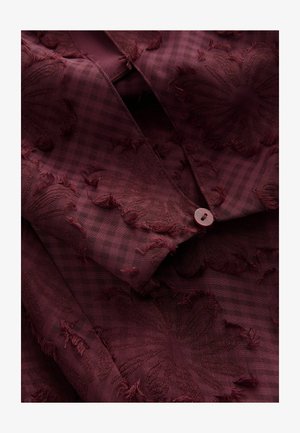 Tela burdeos con un patrón floral texturizado y bordes deshilachados, superpuesto a un sutil diseño de cuadros; incluye un cierre de botón.