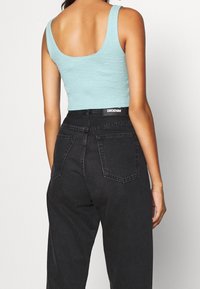 Donna che indossa un top senza maniche blu chiaro e jeans di denim neri a vita alta, vista di spalle su uno sfondo semplice.