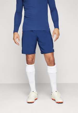 Pantaloni sportivi blu navy realizzati in tessuto leggero, abbinati a calze bianche fino al ginocchio e scarpe da calcio color crema con dettagli arancioni.