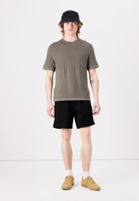 Kortermet olivengrønn ribbestrikket genser; svarte shorts; brune joggesko; svart bøttehatt; brukt med hvite ankelsokker; stående posering.