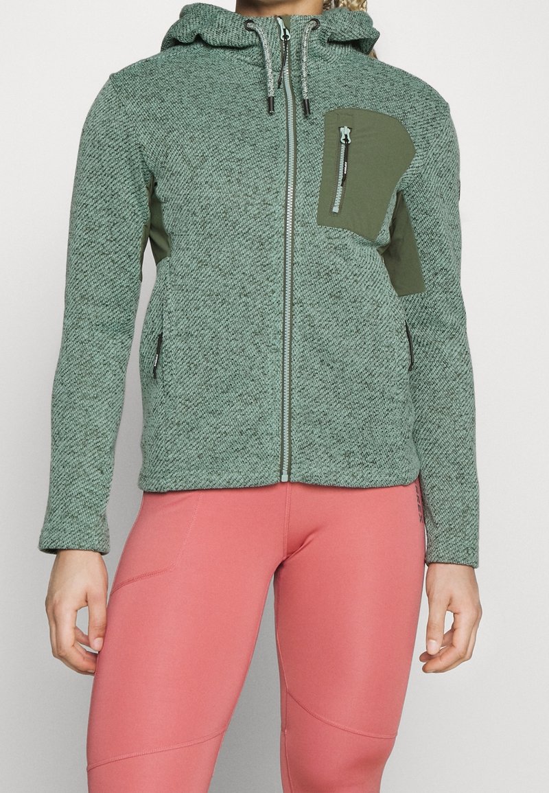 Sweat à capuche zippé vert texturé avec poches zippées poitrine et côtés, porté avec des leggings de sport roses.