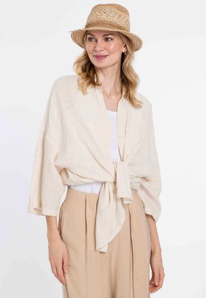 Light beige wrap shirt in textured fabric with three-quarter sleeves, tied at the waist. Paired with cream tank and beige high-waisted trousers.