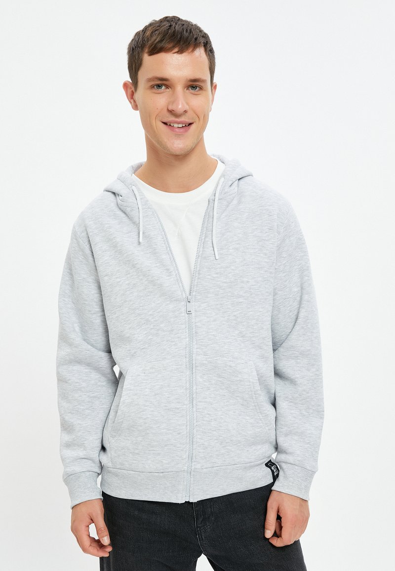 Sudadera con cremallera de color gris claro, con un cierre frontal, dos bolsillos laterales y una capucha con cordón. Fabricada en tejido suave con puños y bajo de canalé.