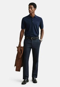 Námořnická polo košile, tmavé kalhoty, černý opasek a černé boty; drží hnědý svetr. Hladká textura, přizpůsobený střih a minimální detaily.