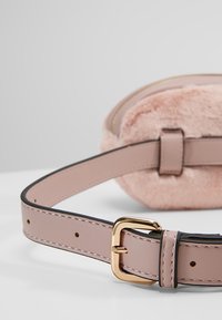 Ceinture rose en cuir synthétique avec boucle dorée attachée à une pochette circulaire rose douce et duveteuse sur une surface blanche.