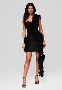 Kvinna klädd i en svart strapless mini-klänning med stora volanger som faller asymmetriskt, tillsammans med svarta klackar med ankeldetalj.